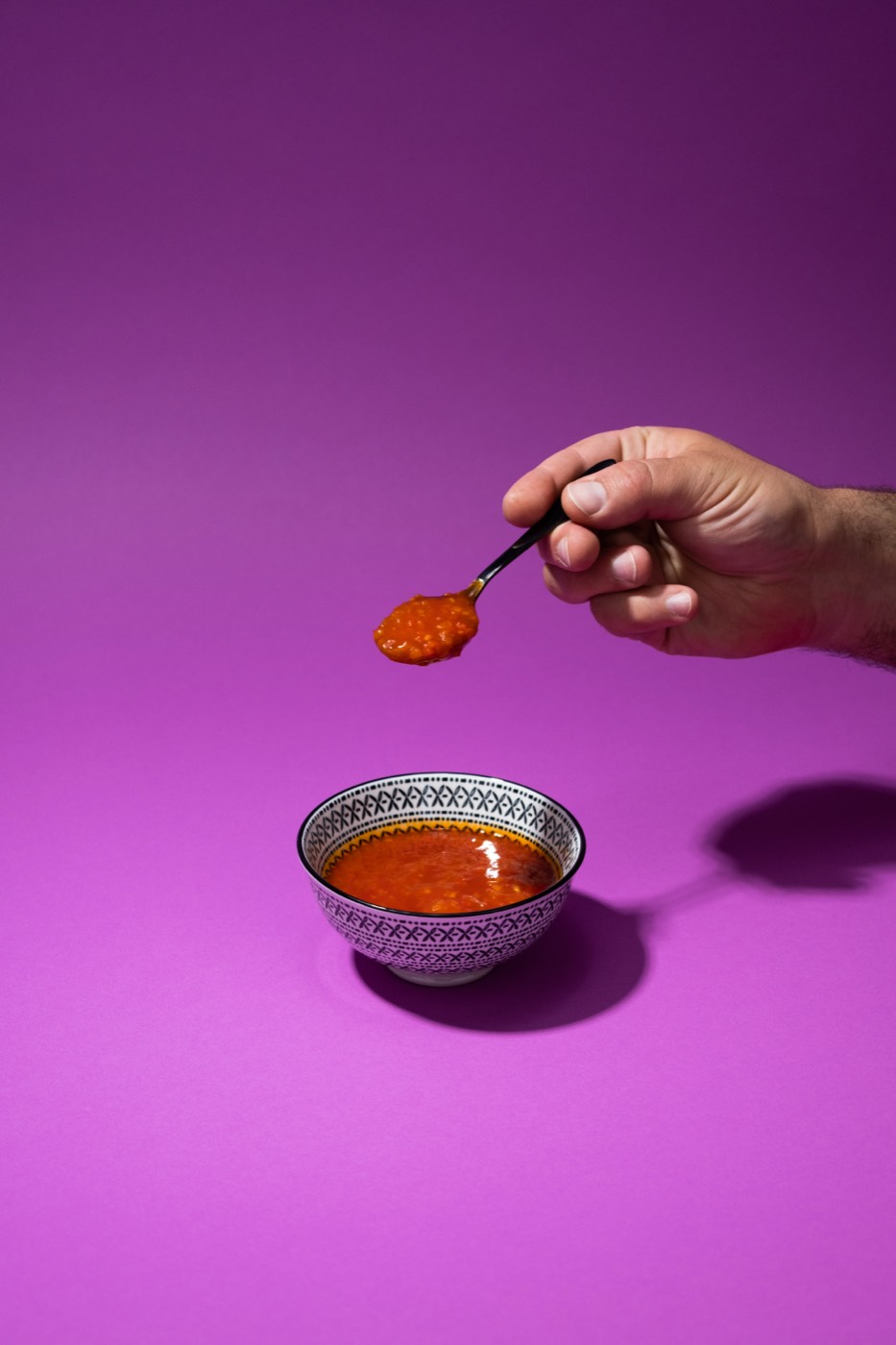 Hand mit Löffel über einer Schale Chilisauce auf violettem Studiohintergrund